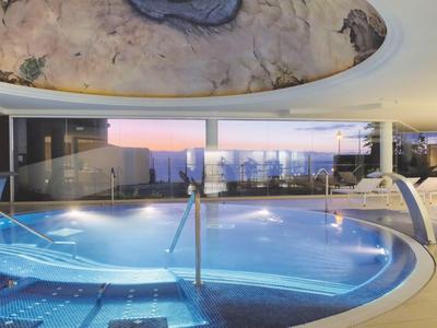 Moderner Innenpool mit blauer Beleuchtung, großen Fenstern und Abendhimmel im Hintergrund.