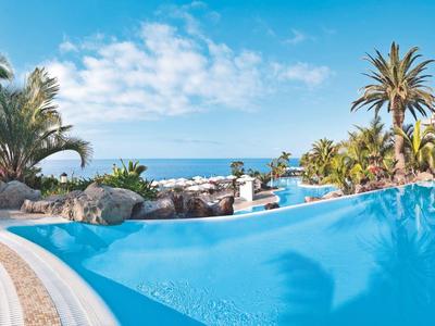 Großer blauer Pool mit Palmen, Sonnenterrasse und Blick aufs Meer unter blauem Himmel.