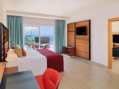 Helles Hotelzimmer mit Doppelbett, TV, Schrank, rotem Sessel und Balkon mit Meerblick.