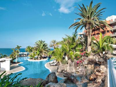 Hotelpool mit Palmen, Felsen und Sonnenschirmen bei strahlend blauem Himmel und Meer im Hintergrund.