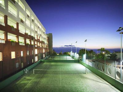 Illuminated tennis court beside a multi-story hotel building at dusk with a view of the sea.