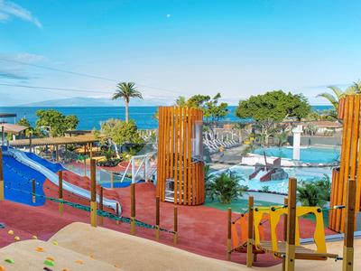 Bunter Kinderspielplatz mit Klettergeräten und Rutschen, Palmen und Meer im Hintergrund.