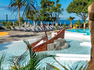 Poollandschaft mit einem Holzboot im flachen Wasser und Liegestühlen am blauen Meer im Hintergrund.
