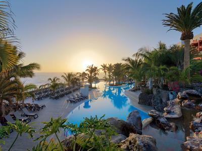 Schwimmbecken mit Palmen und Sonnenuntergang am Meer, umgeben von Liegestühlen und Pflanzen.