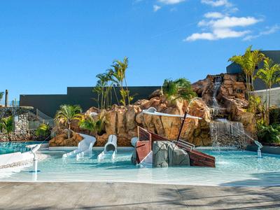 Moderner Pool mit Wasserrutschen, Felsen und Palmen unter blauem Himmel.