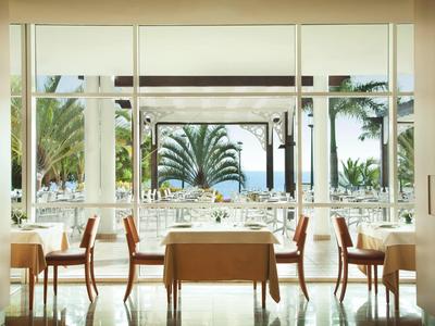 Hell eingerichtetes Restaurant mit Holztischen und Stühlen, Blick auf Palmen und Meer draußen.