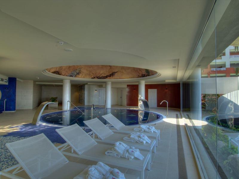 Innenraum mit Whirlpool, weißen Liegestühlen, großen Fenstern und hellem Tageslicht.