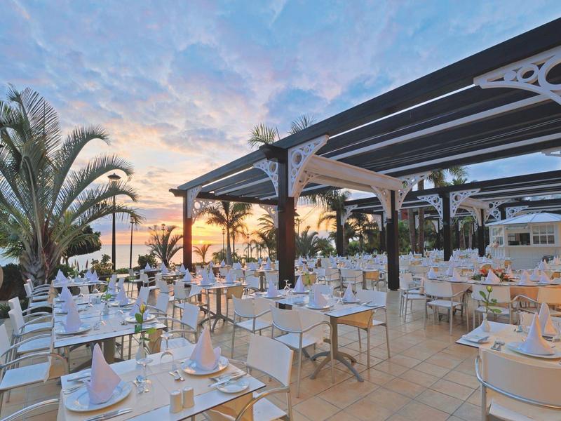 Freiluftrestaurant mit weißen Stühlen und Tischen, Palmen und Sonnenuntergang im Hintergrund.