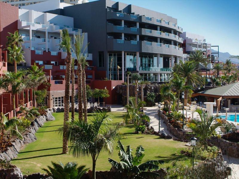 Modernes Hotel mit mehreren Etagen, umgeben von Palmen, grünen Wiesen und einem Poolbereich.