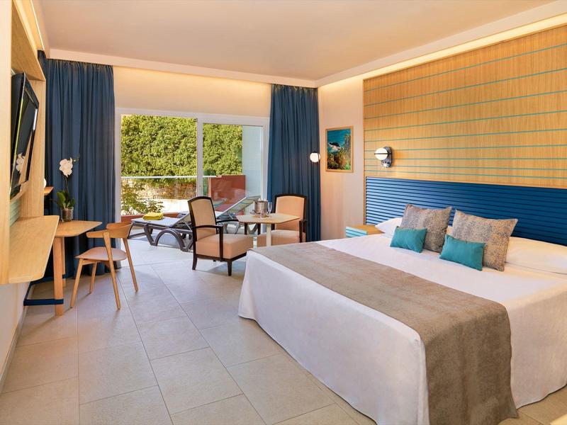 Helles Hotelzimmer mit Doppelbett, blauer Wand, Holzmöbeln und Balkon mit Blick ins Grüne.