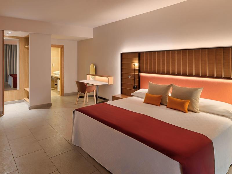 Modernes Hotelzimmer mit großem Bett, rot-weißer Bettdecke, Schreibtisch und indirekter Wandbeleuchtung.