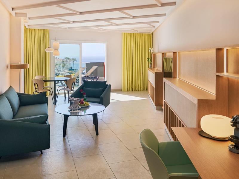 Helles Wohnzimmer mit grünen Möbeln, großen Fenstern und Holzregalen an der Wand.