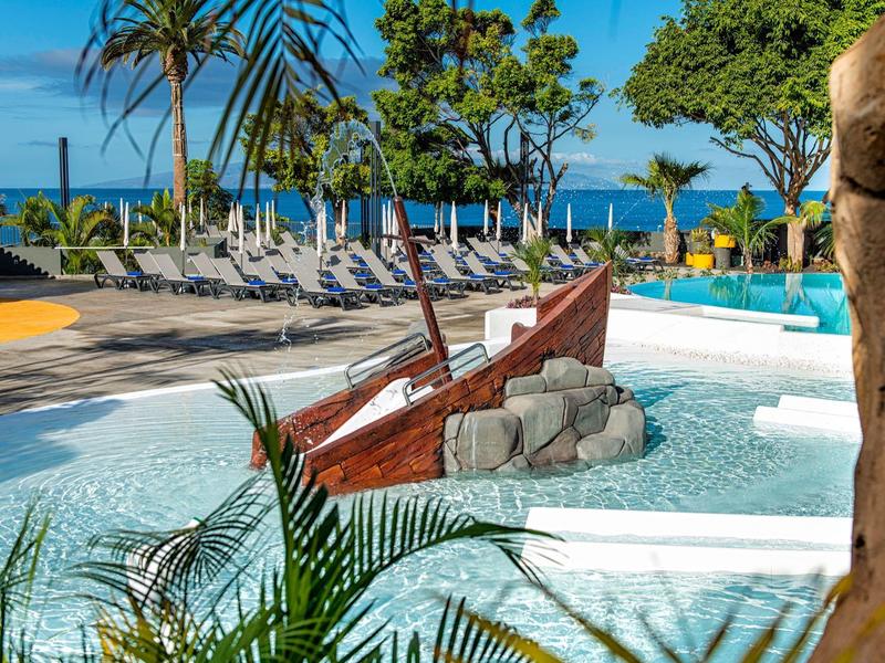 Poollandschaft mit einem Holzboot im flachen Wasser und Liegestühlen am blauen Meer im Hintergrund.