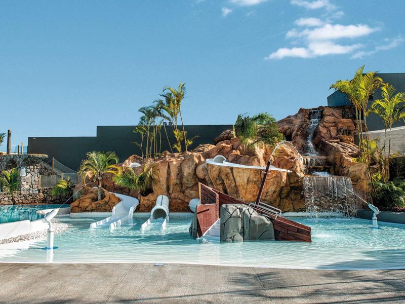 Pool mit Wasserrutschen, Wasserfällen und Palmen vor klarem Himmel und Felsenlandschaft.