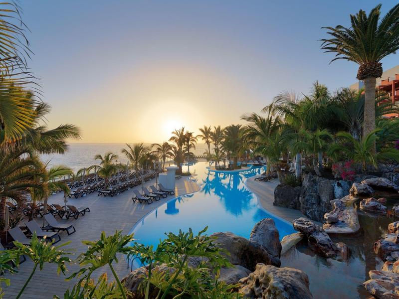 Schwimmbecken mit Palmen und Sonnenuntergang am Meer, umgeben von Liegestühlen und Pflanzen.