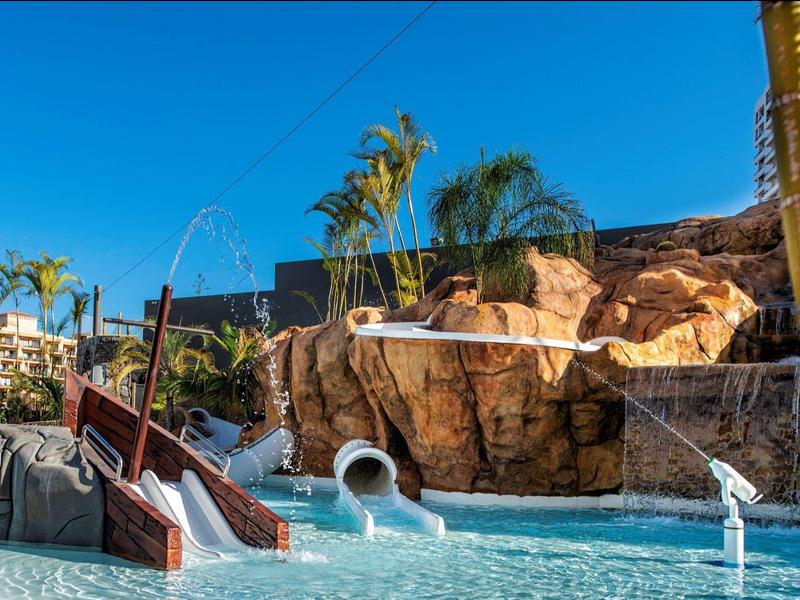 Wasserpark mit Rutschen, künstlichen Felsen, Palmen und klarem blauem Himmel.