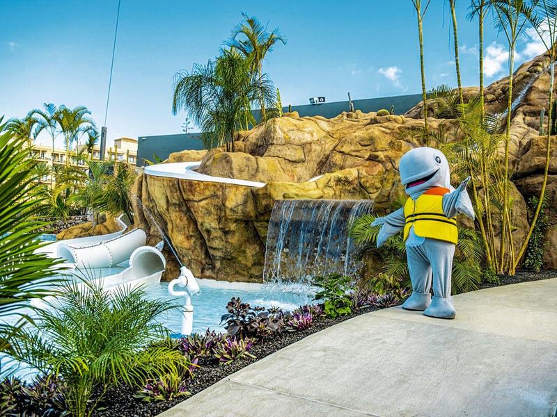 Kind mit buntem Rucksack steht auf Weg vor Wasserfall und Felsen, umgeben von Palmen und Pflanzen.