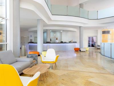 Heller Hotel-Lobbybereich mit modernen Sitzelementen in Gelb und Grau und Rezeption im Hintergrund.