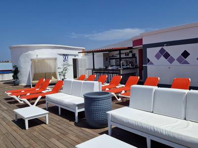 Dachterrasse mit weißen und orangen Sitzmöbeln auf Holzplanken unter blauem Himmel.