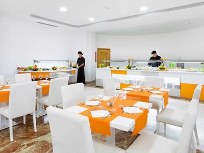 Modernes, helles Restaurant mit weißen Stühlen und orange gedeckten Tischen in einem Hotel.