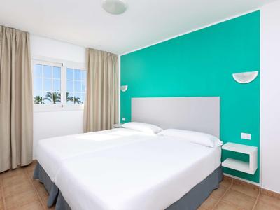 Schlafzimmer mit weißem Bett, türkisfarbener Wand, Fenstern mit Vorhängen und hellbraunem Boden.