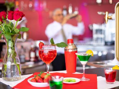 Bar mit roten Getränken, Cocktailshaker, Glasvase mit roten Rosen und Person im Hintergrund.