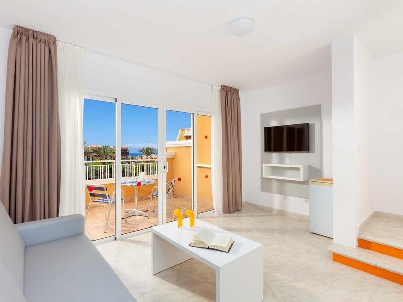 Helles Wohnzimmer mit grauem Sofa, weißen Möbeln, Fernseher und Terrassentür mit Blick auf Balkon.