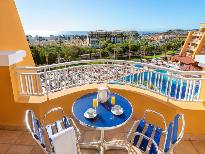 Balkon mit blauem Tisch und zwei Stühlen, Blick auf Pool, Palmen und Stadt im Hintergrund.