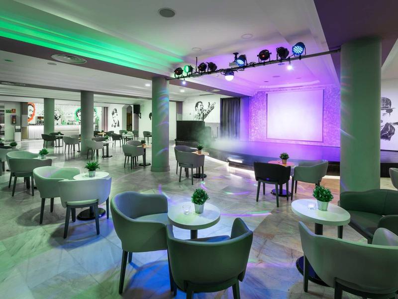 Moderne, helle Lounge mit runden Tischen, grünen Sesseln und bunter Bühnenbeleuchtung.