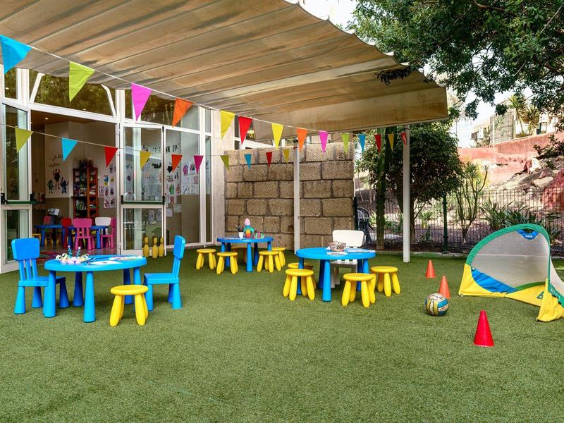 Area giochi esterna colorata con tavoli, sedie, scivolo e decorazioni festive.