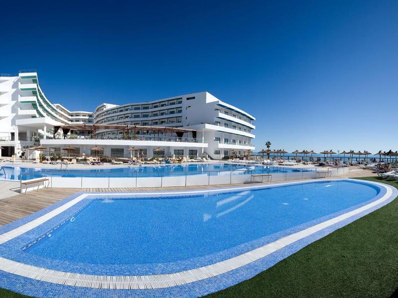 Grande piscina all'aperto con moderno hotel bianco sullo sfondo e cielo azzurro.