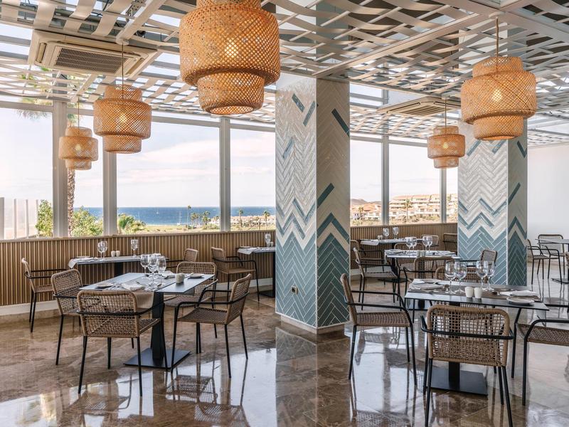 Interno moderno di ristorante con grandi finestre vista mare, tavoli e sedie eleganti.