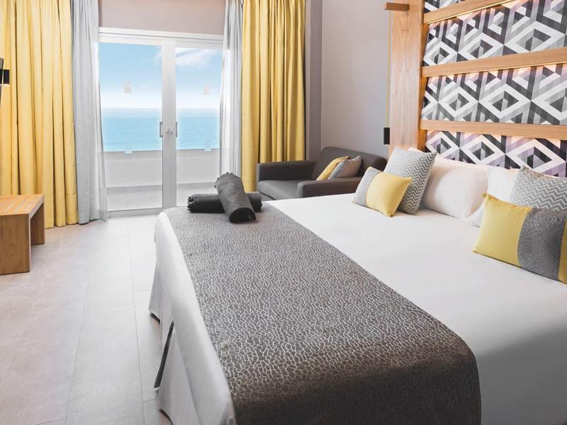 Chambre d'hôtel moderne avec grand lit, canapé, télévision et vue sur la mer par la porte du balcon.