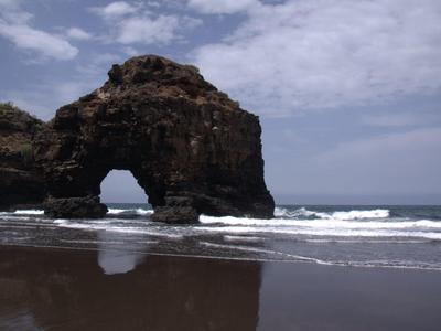 Formazione rocciosa con arco naturale su spiaggia con sabbia scura e mare mosso.