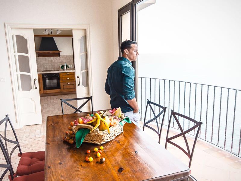 Un uomo è su un balcone e guarda fuori, accanto a un tavolo di legno con frutta.