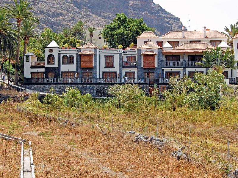 Gebäude mit Terrasse und Grünfläche im Vordergrund, umgeben von Bäumen und Bergen.