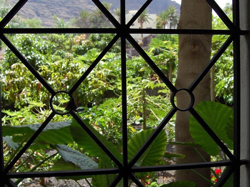 Blick durch ein gitterförmiges Fenster auf grüne Bäume und Berge im Hintergrund.