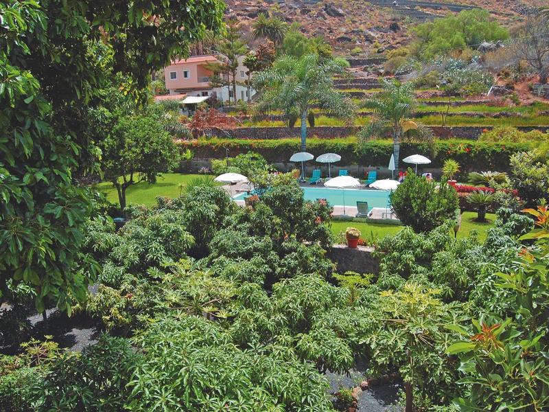 Blick auf einen Garten mit vielen grünen Pflanzen, Sonnenschirmen und Gebäuden in einer hügeligen Landschaft.