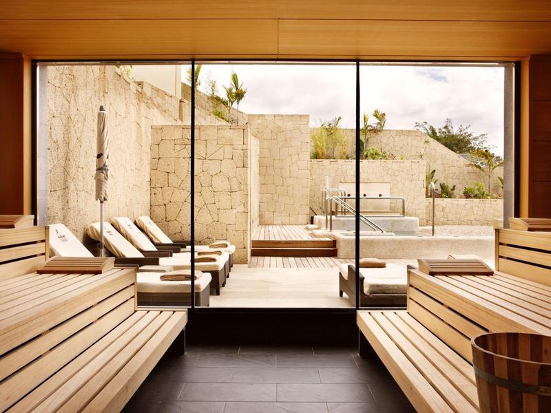 Dřevěná sauna se skleněnými dveřmi vedoucími k venkovnímu bazénu a lehátkům pod oblačnou oblohou.