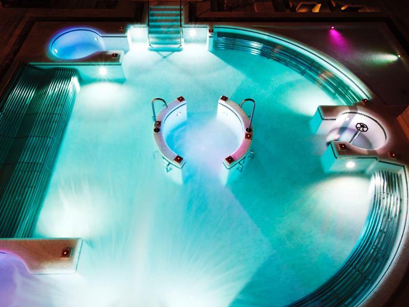 Jasně osvětlený venkovní bazén s zakřivenými schody a posezením zářícím modrými a fialovými odstíny v noci.