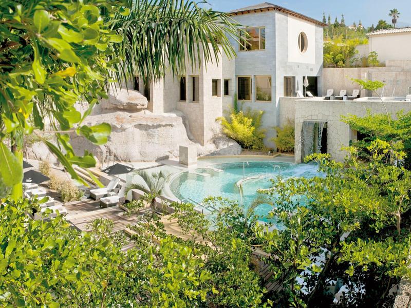 Bílá vila s bazénem obklopená zelení a palmami pod jasnou oblohou.