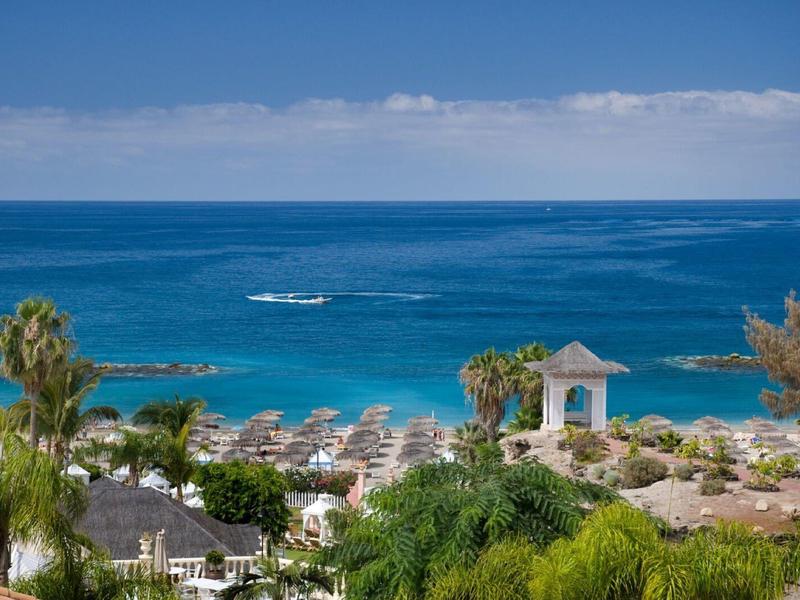 Pobřežní resort s modrým mořem, slunečníky, palmami a malou bílou altánem u pláže.