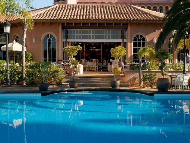 Elegantní venkovní bazén s čistou modrou vodou před hotelem s terakotovou střechou.