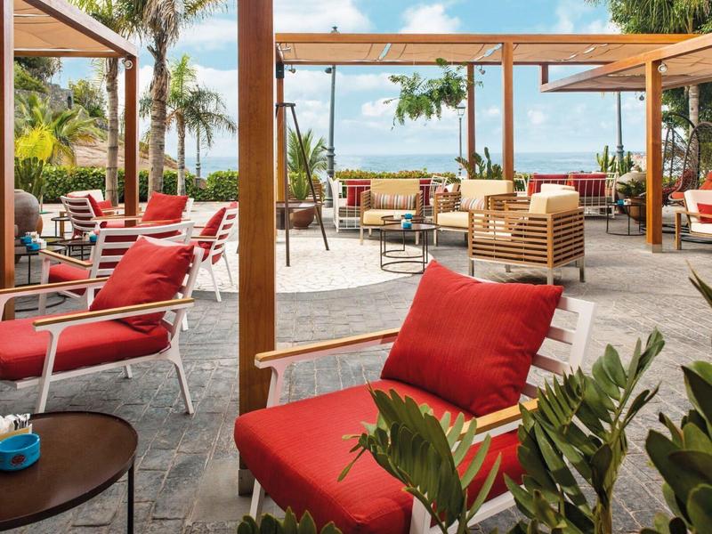 Venkovní posezení s červenými polštáři pod dřevěnými pergolami s výhledem na tropickou zahradu a oblohu.