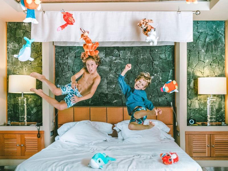 Dva chlapci skáčou na posteli s tropickými rybami zdobícími zelenou zeď za nimi.