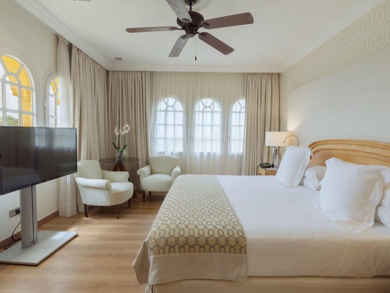 Moderní hotelový pokoj s velkou postelí, televizí, křesly a stropním ventilátorem v světlém prostředí.