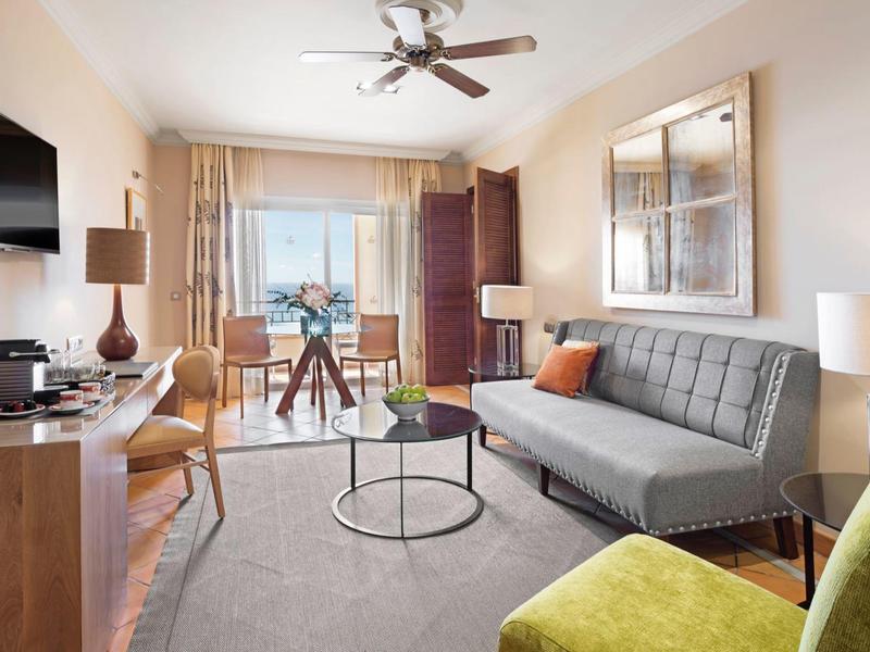 Světý hotelový pokoj se šedou pohovkou, zeleným křeslem, dřevěným nábytkem a velkým oknem s výhledem na město.