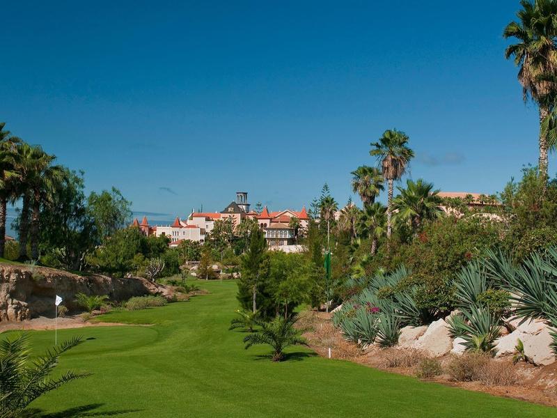 Zelené golfové hřiště lemované palmami pod jasnou modrou oblohou poblíž budov.