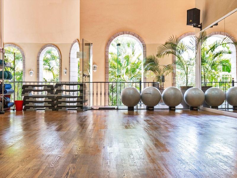 Světelná fitness místnost s dřevěnou podlahou, cvičebními míči, podložkami a velkými obloukovými okny.