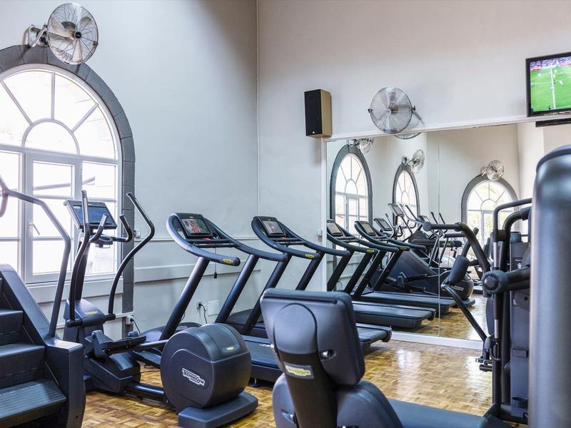 Posilovna s běžeckými pásy, rotopedy a velkými obloukovými okny s nástěnnými ventilátory.